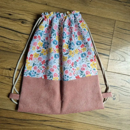 Sac à dos pochon sans rabat, cousu main, en tissu fleuri multicolore avec base en velours côtelé rose et cordons coulissants beiges. Idéal pour les enfants, ce sac combine légèreté, praticité et charme bucolique. Parfait pour la maternelle, les activités ou comme cadeau personnalisé. Présenté à plat sur un parquet bois, il met en valeur un travail artisanal aux finitions soignées.