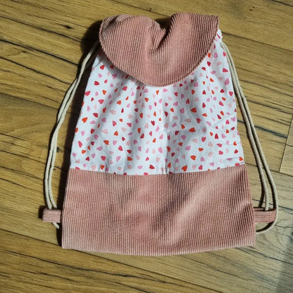 Sac à dos pochon pour enfant confectionné en tissu imprimé de petits cœurs rouges et roses sur fond blanc, avec base et rabat en velours côtelé rose. Fermeture par cordon coulissant beige. Accessoire fait main au style doux et romantique, idéal pour la crèche, la maternelle ou les sorties. Posé sur un sol en bois, ce sac à dos associe praticité, confort et design tendance pour un usage quotidien.