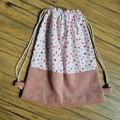 Sac à dos pochon enfant cousu main, réalisé en tissu blanc à motifs cœurs rouges et roses, avec une base en velours côtelé rose. Fermeture par cordon coulissant beige. Design doux et pratique, idéal pour la maternelle, les sorties ou les activités sportives. Ce modèle sans rabat allie légèreté, solidité et style enfantin, avec une finition soignée mise en valeur sur un parquet en bois naturel.