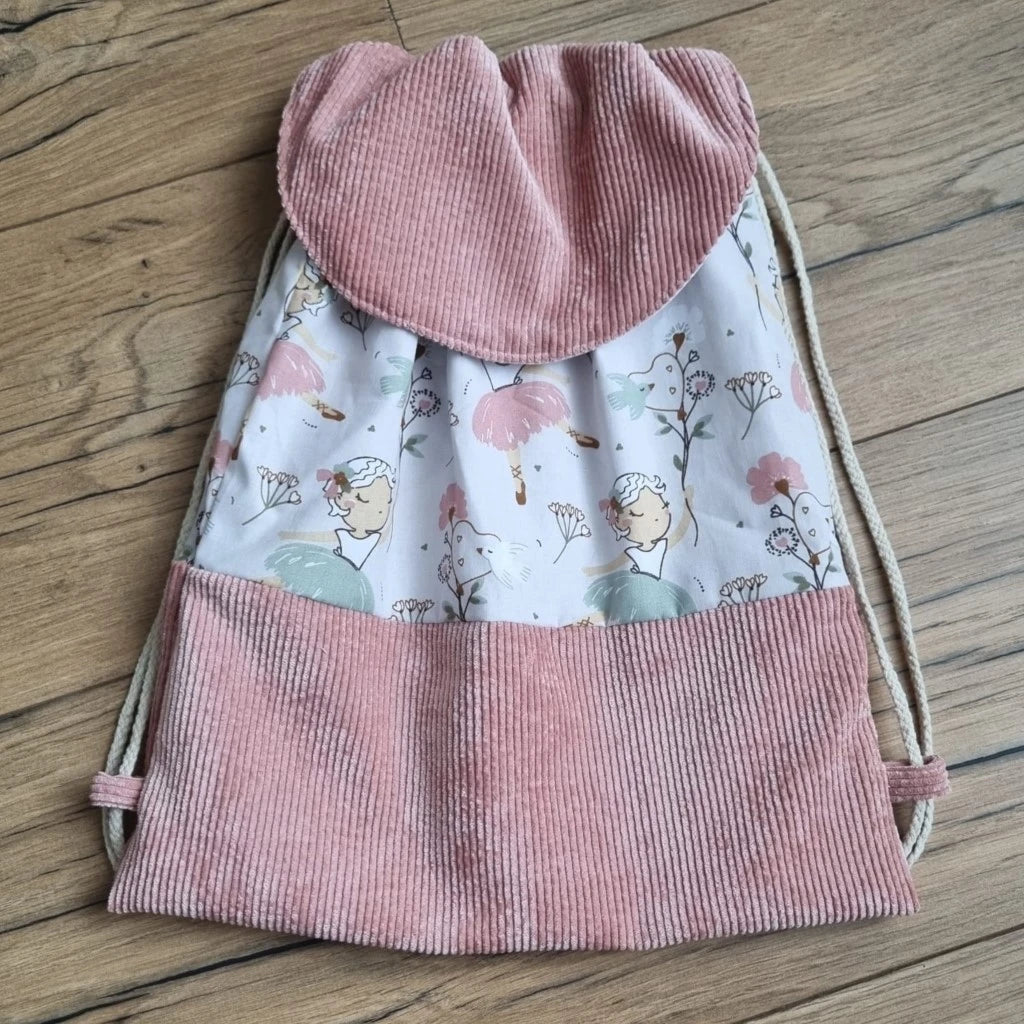 Sac à dos au format pochon cousu dans un tissu coton imprimé au motif danseuse. La base du sac est en velours côtelé rose. Le sac se ferme grâce à deux cordelettes de part et d'autre en coton blanc. Le sac dispose d'un rabat arrondi en velours côtelé rose identique à celui de la base du sac.