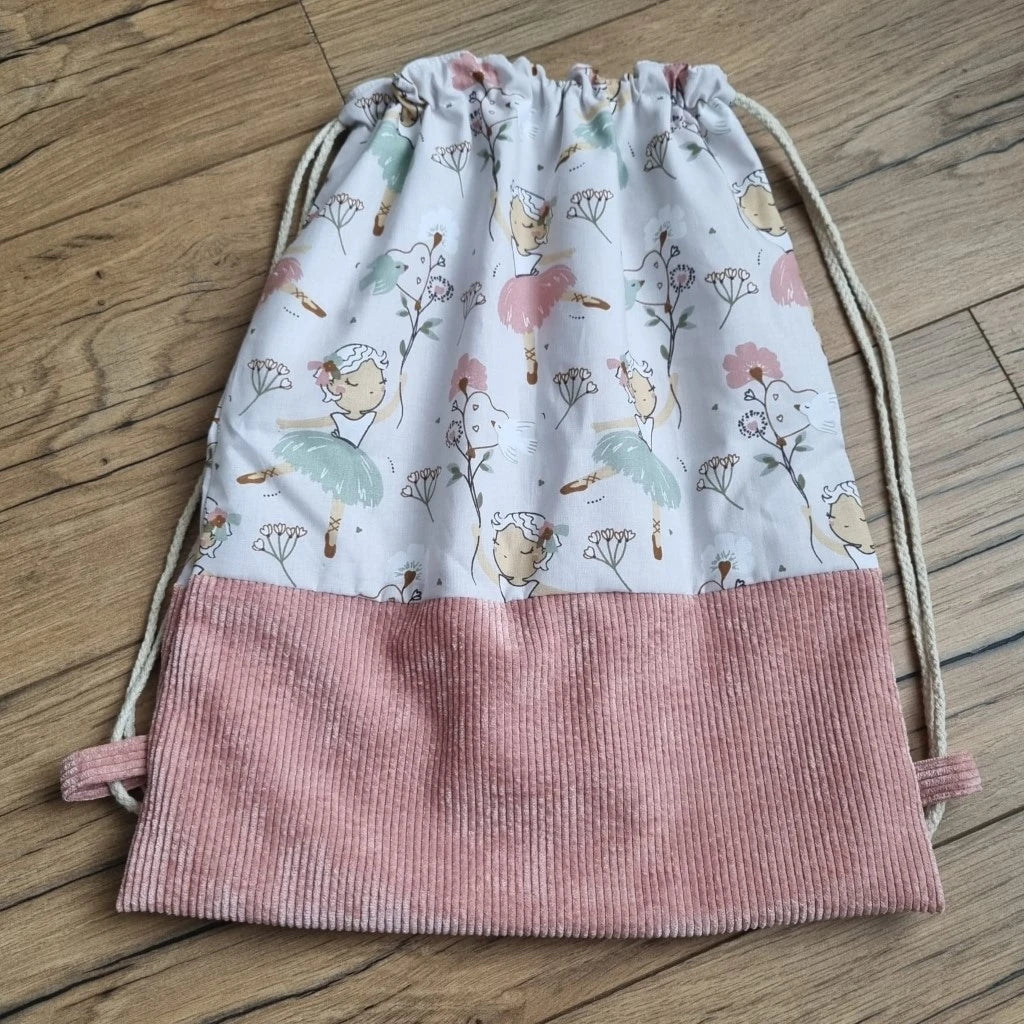 Sac à dos au format pochon cousu dans un tissu coton imprimé à motif danseuse. La base du sac est en velours côtelé rose. Le sac se ferme grâce à deux cordelettes de part et d'autre en coton blanc.