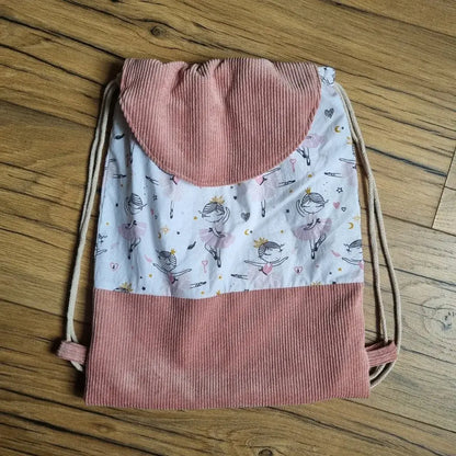 Sac à dos pochon fait main pour enfant, conçu en velours côtelé rose et tissu blanc à motifs de petites danseuses classiques. Fermeture par cordon coulissant et rabat arrondi pour un style soigné et original. Idéal pour l’école, les activités de danse ou les sorties. Le sac est posé à plat sur un parquet en bois, mettant en valeur ses finitions et son design ludique.