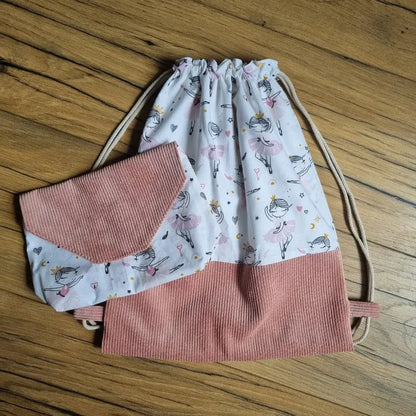 Ensemble assorti composé d’un sac à dos pochon sans rabat et d’une pochette, cousus main en velours côtelé rose et tissu blanc imprimé de petites danseuses. Le sac se ferme par cordon coulissant et la pochette par rabat en triangle. Idéal pour l’école, la danse ou les loisirs. Accessoires posés sur un sol en bois naturel, mettant en valeur leur design doux et enfantin.