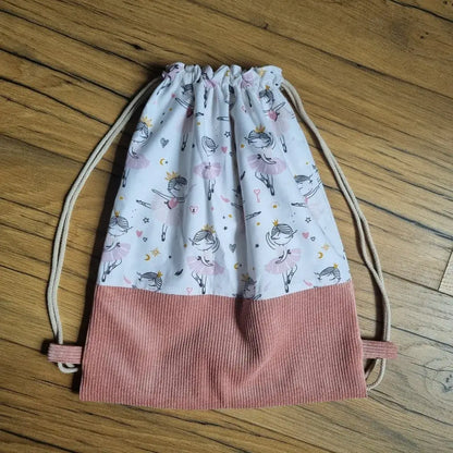 Sac à dos pochon pour enfant confectionné à la main, avec base en velours côtelé rose et tissu blanc imprimé de petites danseuses classiques en tutu rose. Fermeture par cordon coulissant beige. Idéal pour les activités de danse, l’école ou les sorties. Le sac est posé à plat sur un sol en bois naturel, mettant en valeur son design délicat et ses finitions soignées.