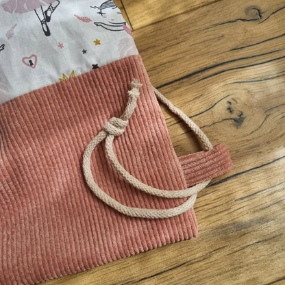 Gros plan sur la base d’un sac à dos pochon cousu main, en velours côtelé rose saumon avec cordon de serrage en coton beige. Le tissu du sac est imprimé de petites danseuses en tutu rose. L’image met en valeur la qualité des finitions, le détail des coutures et la solidité du système de fermeture, le tout sur un fond en bois naturel.