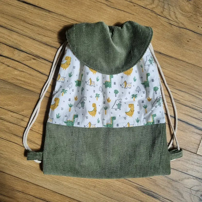 Sac à dos pochon pour enfant fabriqué à la main, avec rabat en velours côtelé vert kaki et tissu imprimé dinosaures colorés sur fond blanc. Fermeture par cordons coulissants beiges. Idéal pour la maternelle ou les sorties, ce sac léger et pratique allie originalité et confort. Motifs ludiques de dinosaures jaunes et verts pour plaire aux petits explorateurs. Accessoire artisanal parfait pour un cadeau personnalisé.