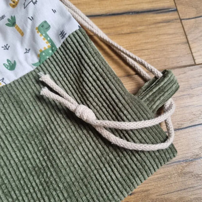 Zoom sur la base en velours côtelé vert kaki et les cordelettes beiges d’un sac à dos pochon enfant. Détail de couture soignée et finitions robustes, idéal pour un usage quotidien. Le tissu à motifs dinosaures colorés complète ce sac pratique et original, parfait pour la maternelle ou les sorties. Fabrication artisanale mettant en valeur la qualité des matériaux.
