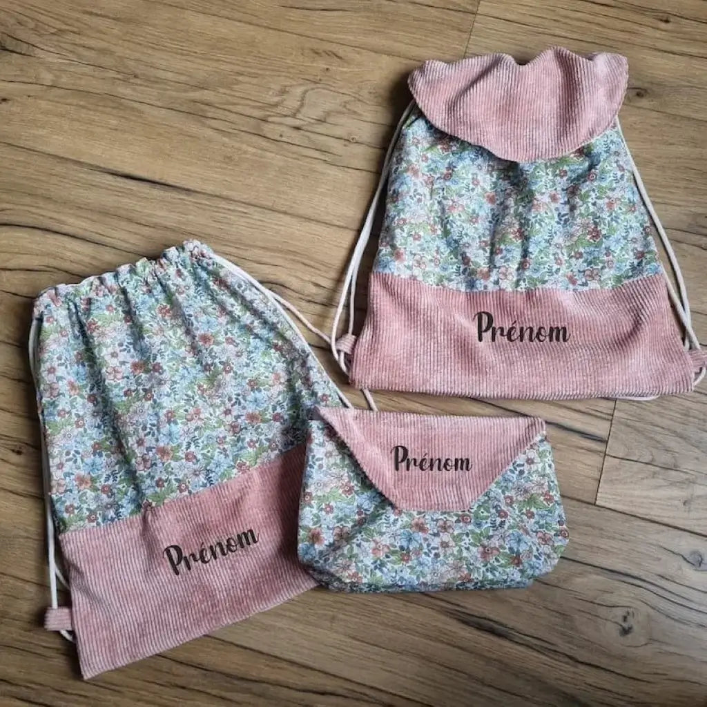 Ensemble assorti de deux sacs à dos pochons et d'une pochette imperméable au motif fleurs. Le sac à dos de gauche est sans rabat. Celui de droite dispose d'un rabat de couleur rose. Les trois articles sont personnalisables avec un prénom floqué sur le rabat ou la base en velours côtelé.