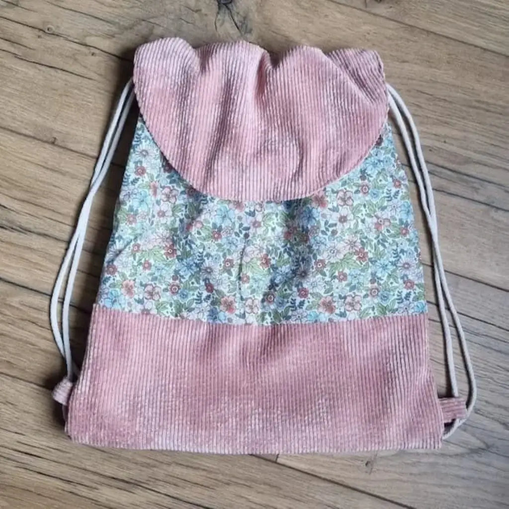 Sac à dos au format pochon cousu dans un tissu coton imprimé à motif fleurs. La base du sac est en velours côtelé rose. Le sac se ferme grâce à deux cordelettes de part et d'autre en coton blanc. Le sac dispose d'un rabat arrondi en velours côtelé rose identique à celui de la base du sac.