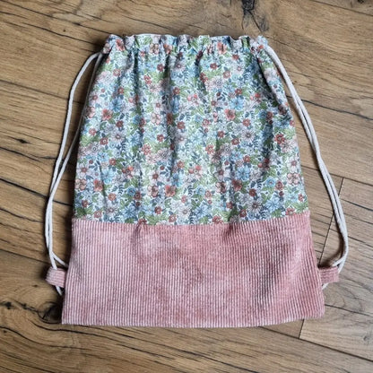 Sac à dos au format pochon cousu dans un tissu coton imprimé à motif fleurs. La base du sac est en velours côtelé rose. Le sac se ferme grâce à deux cordelettes de part et d'autre en coton blanc.