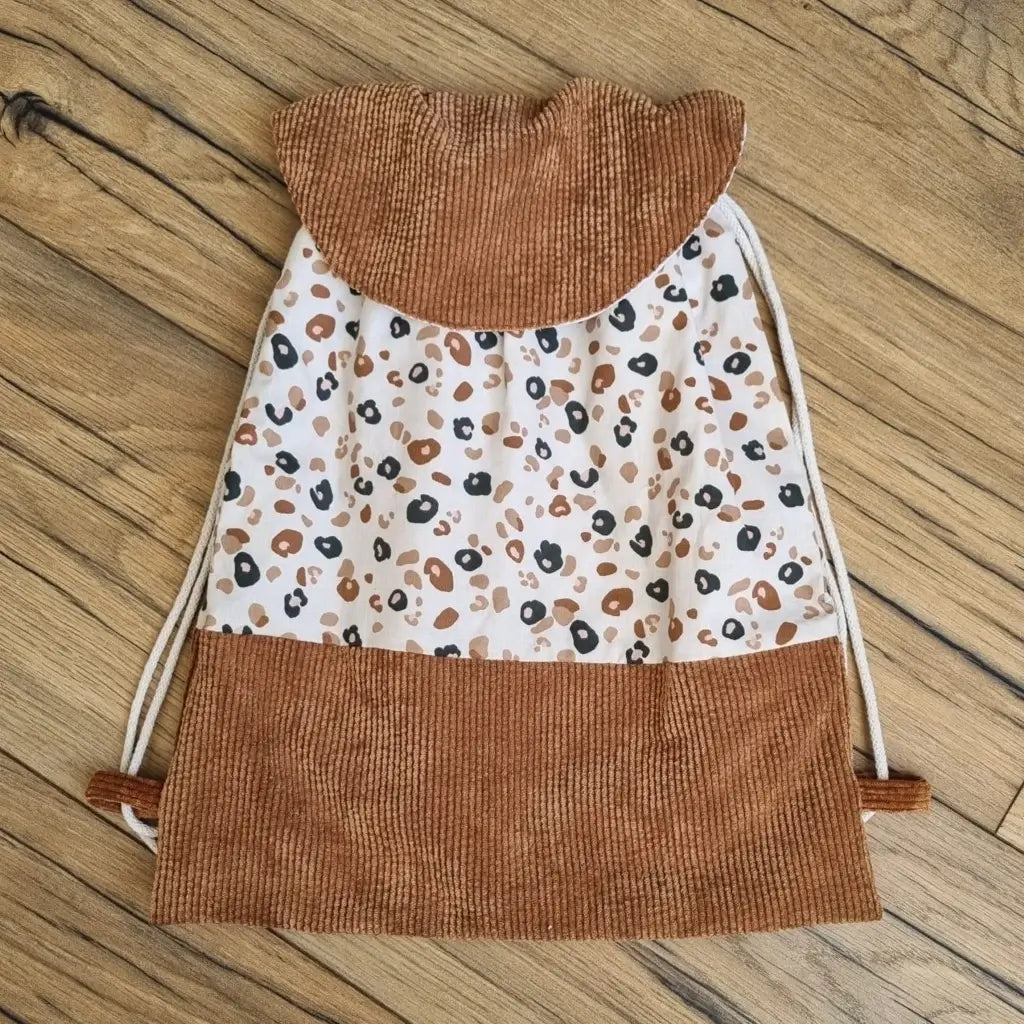 Sac à dos au format pochon cousu dans un tissu coton imprimé à motif léopard. La base du sac est en velours côtelé marron. Le sac se ferme grâce à deux cordelettes de part et d'autre en coton blanc. Le sac dispose d'un rabat arrondi en velours côtelé marron identique à celui de la base du sac.