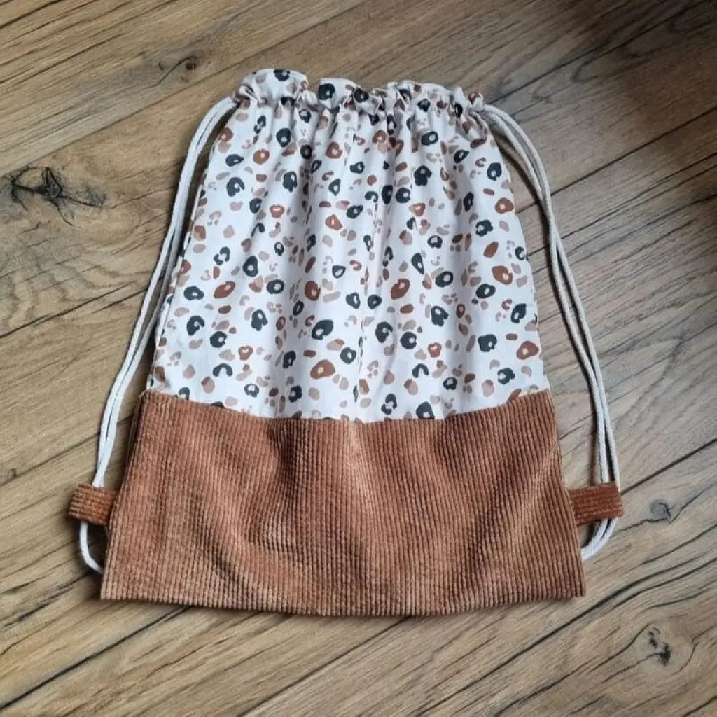 Sac à dos au format pochon cousu dans un tissu coton imprimé à motif léopard. La base du sac est en velours côtelé marron. Le sac se ferme grâce à deux cordelettes de part et d'autre en coton blanc.