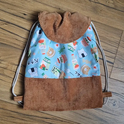 Sac à dos pochon pour enfant fait main, avec rabat en velours côtelé marron et tissu bleu à motifs kawaii représentant des animaux mignons et des aliments japonais (sushis, bubble tea, lait, fraises). Fermeture par cordons coulissants. Idéal pour l’école, la crèche ou les sorties.