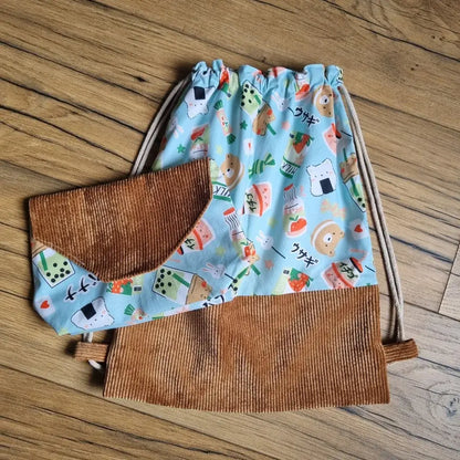 Sac à dos pochon pour enfant assorti à une pochette, confectionnés en tissu bleu à motifs kawaii (bubble tea, animaux, sushis, fraises) et velours côtelé marron. Modèle sans rabat, à fermeture par cordons coulissants. Ensemble fait main idéal pour l’école, la crèche ou les sorties.