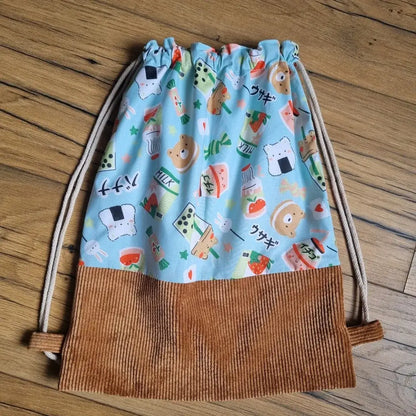 Sac à dos pochon pour enfant, cousu main, sans rabat, avec base en velours côtelé marron et tissu bleu à motifs kawaii représentant des animaux mignons, bubble tea, fraises, briques de lait et sushis. Fermeture par cordons coulissants. Idéal pour l’école, la crèche ou les activités.