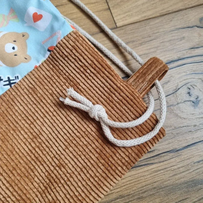 Zoom sur la base en velours côtelé marron d’un sac à dos pochon pour enfant, avec cordon de serrage en coton écru noué et tissu bleu à motifs kawaii visibles en haut (ours, cœur, écriture japonaise). Détail soigné de fabrication artisanale.