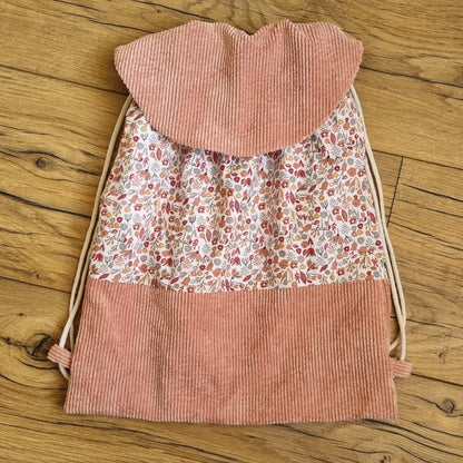 Sac à dos format pochon. Le corps du sac est composé d'un tissu au motif liberty. La base et le rabat du sac sont en velours côtelé rose. Deux cordelettes en coton blanc, de part et d'autre du sac, permettent sa fermeture.