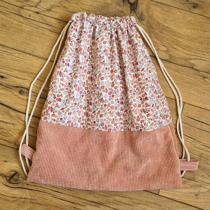 Sac à dos format pochon. Le corps du sac est composé d'un tissu au motif liberty. La base du sac est en velours côtelé rose. Deux cordelettes en coton blanc, de part et d'autre du sac, permettent sa fermeture.