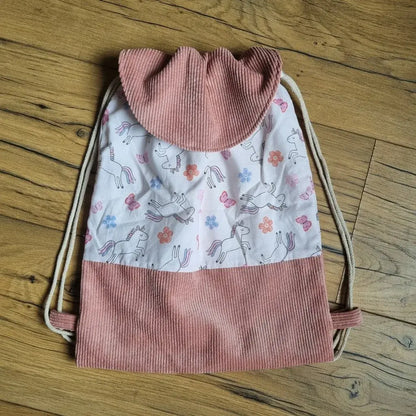 Sac à dos pochon pour enfant, fait main en tissu imprimé licornes, fleurs et papillons sur fond rose pâle, avec rabat et base en velours côtelé rose. Fermeture par cordons écrus, idéal pour la maternelle, les sorties ou en cadeau personnalisé. Présenté à plat sur un sol en bois.