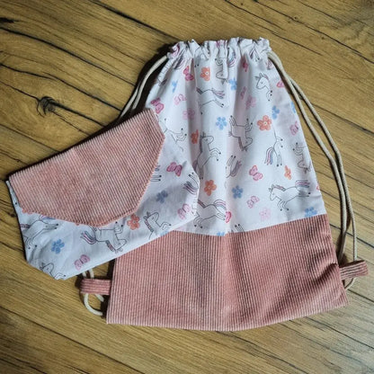 Sac à dos pochon sans rabat en tissu imprimé licornes, fleurs et papillons colorés sur fond rose pâle, avec base en velours côtelé rose. Accompagné d’une pochette assortie posée sur le sac. Ensemble fait main, idéal pour la maternelle, les loisirs ou en cadeau personnalisé, présenté à plat sur un sol en bois.