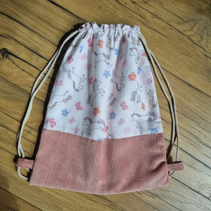 Sac à dos pochon sans rabat, fait main, en tissu imprimé licornes, fleurs et papillons colorés sur fond rose pâle, avec base en velours côtelé rose et cordons écrus servant de bretelles. Accessoire léger et pratique, idéal pour la maternelle ou les activités des enfants, posé à plat sur un sol en bois.