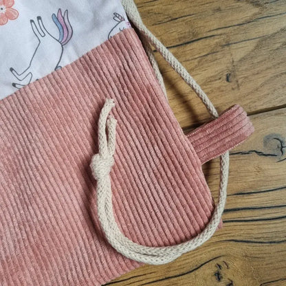 Gros plan sur la base d’un sac à dos pochon enfant, en velours côtelé rose avec cordon écru noué. Partie supérieure en tissu imprimé de licornes, fleurs et papillons colorés sur fond rose pâle. Détail du passe-cordon assorti, photo prise sur un sol en bois clair.