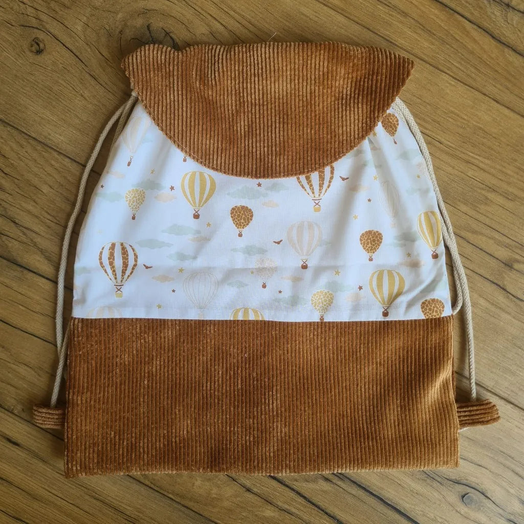 Sac à dos au format pochon cousu dans un tissu coton imprimé au motif montgolfière. La base du sac est en velours côtelé marron. Le sac se ferme grâce à deux cordelettes de part et d'autre en coton blanc. Le sac dispose d'un rabat arrondi en velours côtelé marron identique à celui de la base du sac.