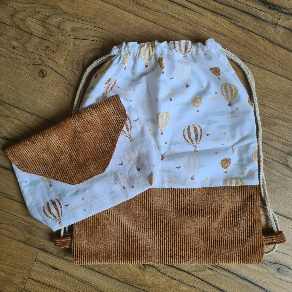 Ensemble assorti d'un sac à dos pochon et d'une pochette imperméable au motif montgolfière. A droite, le sac à dos au format pochon cousu dans un tissu imprimé au motif montgolfière avec une base en velours côtelé marron. Ce sac se ferme grâce à deux cordelettes en coton blanc de part et d'autre du sac. A gauche, une petite pochette imperméable cousu dans le même tissu au motif montgolfière et disposant d'un rabat en velours côtelé marron également.