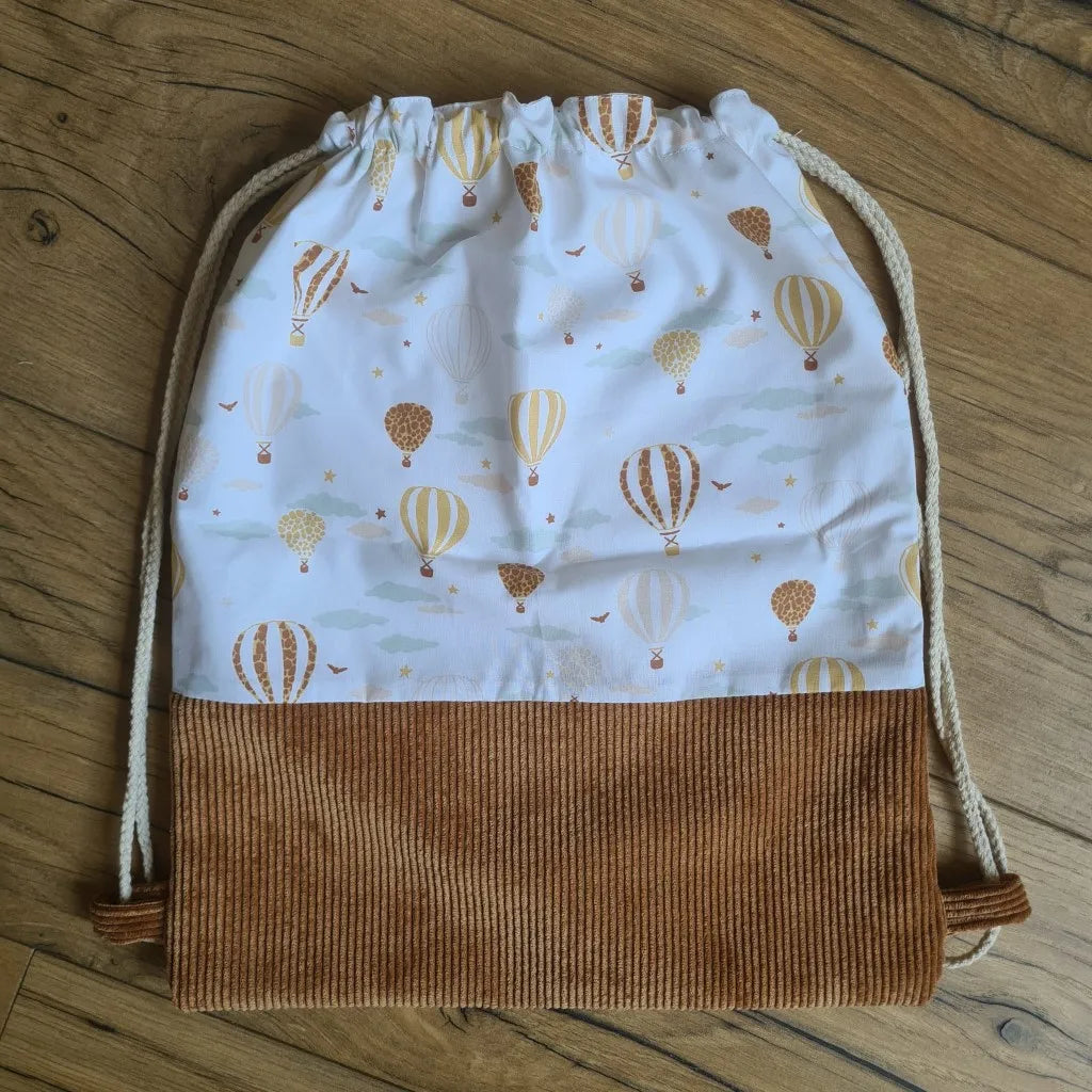 Sac à dos au format pochon cousu dans un tissu coton imprimé à motif montgolfière. La base du sac est en velours côtelé marron. Le sac se ferme grâce à deux cordelettes de part et d'autre en coton blanc.