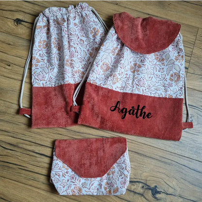 Ensemble fait main composé de deux sacs à dos pochon en tissu à motif oriental rose et terracotta, avec base en velours côtelé terracotta, cordons écrus et rabat assorti sur l’un des sacs, personnalisé avec le prénom Agathe. Pochette coordonnée assortie posée en bas, le tout présenté à plat sur un sol en bois.