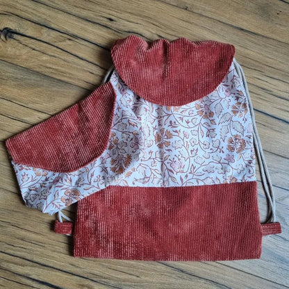 Sac à dos pochon artisanal en tissu fleuri style oriental avec rabat et base en velours côtelé terracotta, accompagné d’une pochette assortie posée sur le sac. Ensemble fait main, idéal pour la maternelle, les sorties ou comme idée cadeau personnalisé, présenté sur un sol en bois.