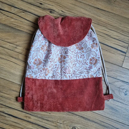 Sac à dos pochon artisanal en tissu à motif floral oriental dans les tons terracotta et rose, avec rabat et base en velours côtelé terracotta. Fermeture par cordon écru et finitions soignées, idéal pour enfant ou adulte, présenté à plat sur un sol en bois.