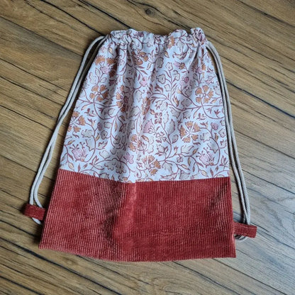 Sac à dos pochon sans rabat, confectionné à la main avec tissu en coton à motif floral oriental dans les tons terracotta et rose, et base en velours côtelé terracotta. Fermeture par cordons écrus servant aussi de bretelles. Accessoire léger et pratique, posé à plat sur un sol en bois.