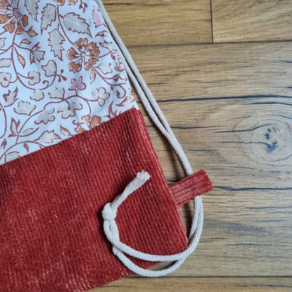 Zoom sur la base d’un sac à dos pochon fait main, en velours côtelé rouge brique avec cordon écru noué. Partie supérieure en coton à motif floral oriental rose et terracotta. Détail de la couture et du passe-cordon visible, posé à plat sur un sol en bois clair.