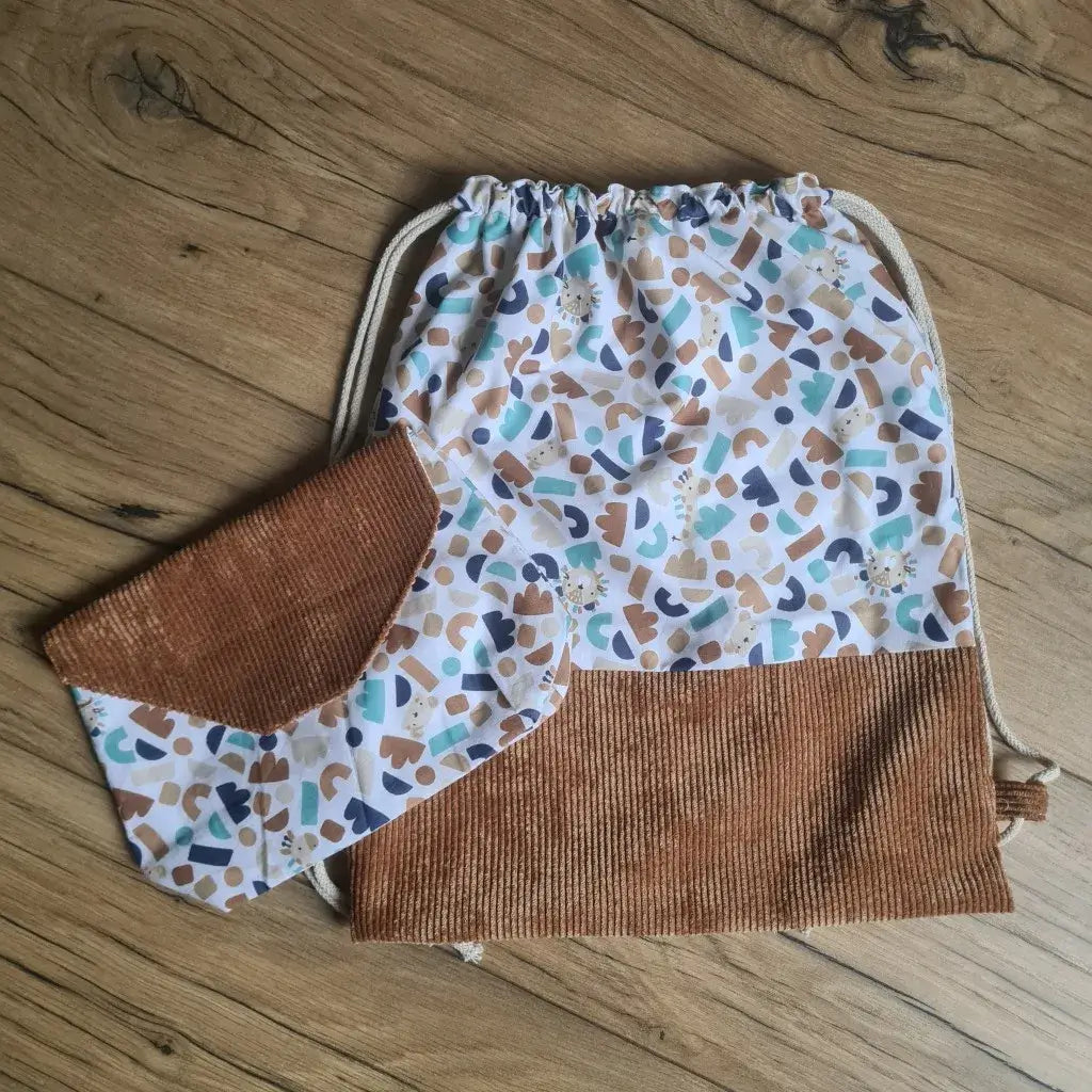 Ensemble assorti d'un sac à dos pochon et d'une pochette imperméable au motif ptiwi. A droite, le sac à dos au format pochon cousu dans un tissu imprimé au motif ptiwi avec une base en velours côtelé marron. Ce sac se ferme grâce à deux cordelettes en coton blanc de part et d'autre du sac. A gauche, une petite pochette imperméable cousu dans le même tissu au motif ptiwi et disposant d'un rabat en velours côtelé marron également.