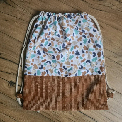 Sac à dos au format pochon cousu dans un tissu coton imprimé à motif ptiwi. La base du sac est en velours côtelé marron. Le sac se ferme grâce à deux cordelettes de part et d'autre en coton blanc.