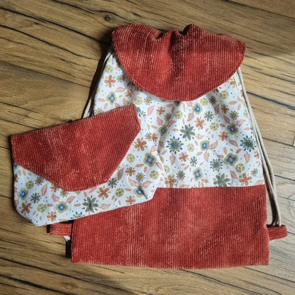 Sac à dos pochon et pochette assortie faits main, en velours côtelé terracotta et tissu fleuri motif Rozia. Le sac dispose d’un rabat arrondi et de bretelles en cordon écru. La pochette présente un rabat en pointe dans le même tissu. Ensemble coordonné posé sur un sol en bois, idéal pour enfant ou pour offrir en cadeau personnalisé.