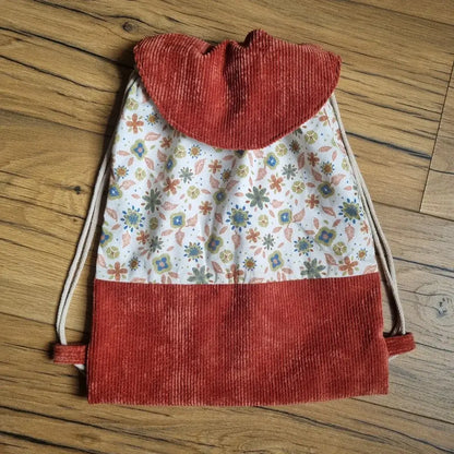 Sac à dos pochon artisanal pour enfant, confectionné en velours côtelé terracotta et tissu coton fleuri motif Rozia. Le rabat arrondi est assorti au bas du sac. Les bretelles en cordon écru permettent un port confortable. Posé sur un sol en bois, ce sac fait main est idéal pour la maternelle, les sorties ou les cadeaux personnalisés.