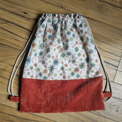 Sac à dos pochon en tissu avec bas en velours côtelé terracotta et haut en coton imprimé floral dans les tons pastel, doté de cordons beiges servant de bretelles, posé à plat sur un sol en bois. Idéal pour un usage quotidien, léger et fait main.
