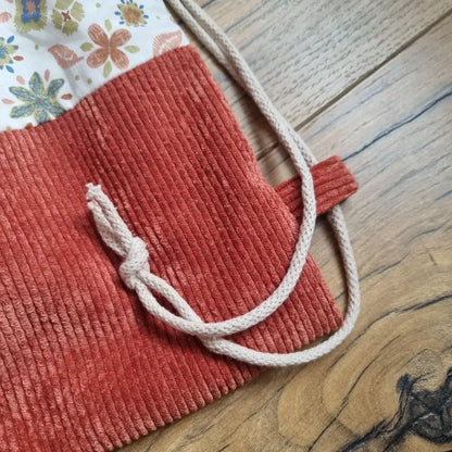 Gros plan sur la base d’un sac à dos pochon fait main, en velours côtelé terracotta avec cordon de serrage écru noué, et tissu supérieur à motif floral. Zoom sur les détails de couture et la finition du passe-cordon, posé sur un sol en bois.