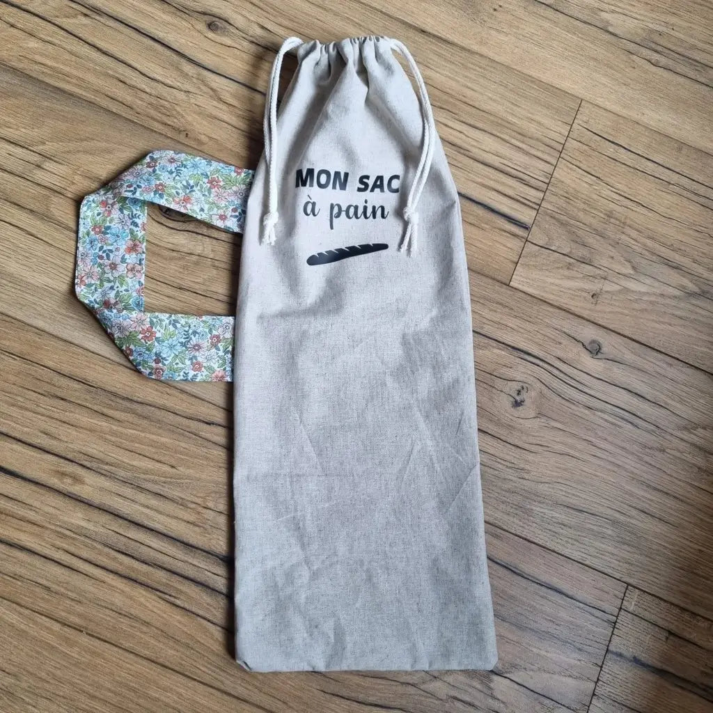 Sac à pain en coton au format baguette se fermant par deux petites cordelettes blanches avec une sangle au motif fleuri et l'inscription "Mon sac à pain" et le logo d'une baguette floqués au centre sur la partie supérieure haute.