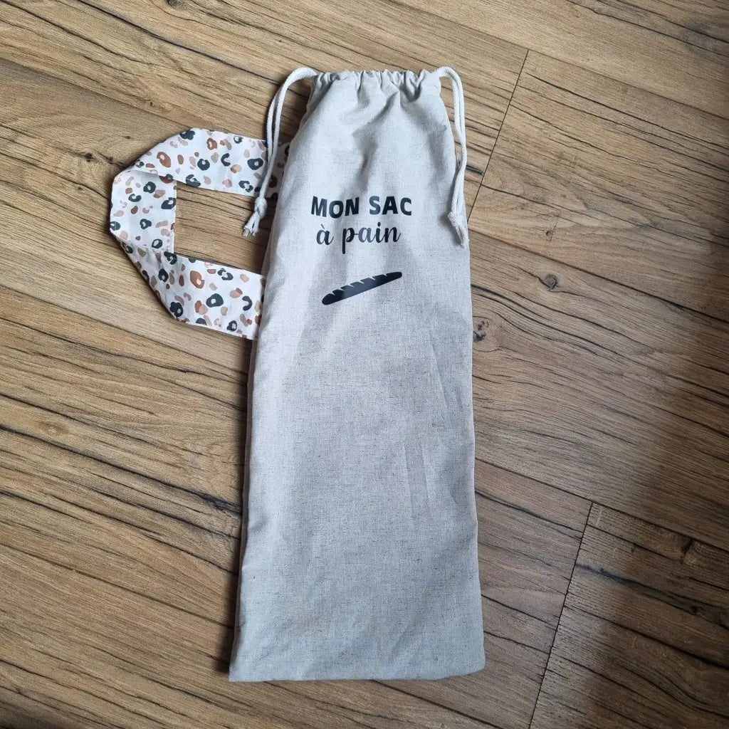 Sac à pain en coton au format baguette se fermant par deux petites cordelettes blanches avec une sangle au motif léopard et l'inscription "Mon sac à pain" et le logo d'une baguette floqués au centre sur la partie supérieure haute.