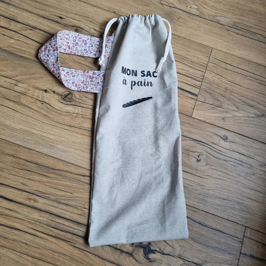 Sac à pain en coton au format baguette se fermant par deux petites cordelettes blanches avec une sangle au motif Liberty et l'inscription "Mon sac à pain" et le logo d'une baguette floqués au centre sur la partie supérieure haute.