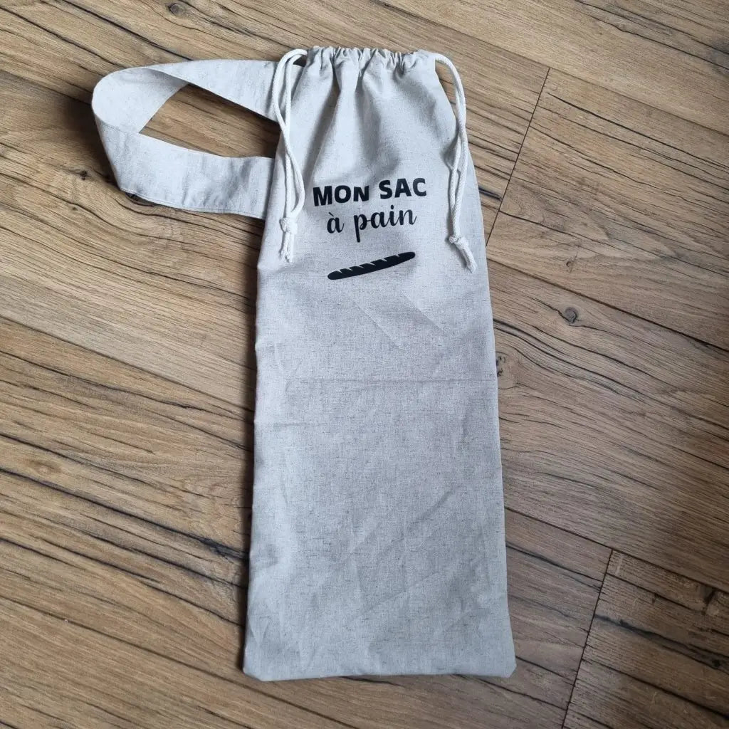 Sac à pain en coton au format baguette se fermant par deux petites cordelettes blanches avec une sangle au motif uni du même coloris que l'ensemble et l'inscription "Mon sac à pain" et le logo d'une baguette floqués au centre sur la partie supérieure haute.