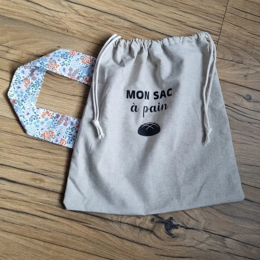 Sac à pain en coton au format miche se fermant par deux petites cordelettes blanches avec une sangle au motif fleuri et l'inscription "Mon sac à pain" et le logo d'une miche de pain floqués au centre sur la partie supérieure haute.