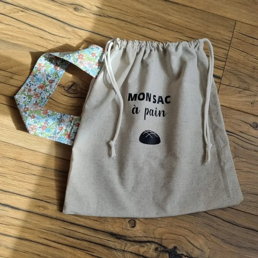 Sac à pain en coton au format miche se fermant par deux petites cordelettes blanches avec une sangle au motif fleuri et l'inscription "Mon sac à pain" et le logo d'une miche de pain floqués au centre sur la partie supérieure haute.