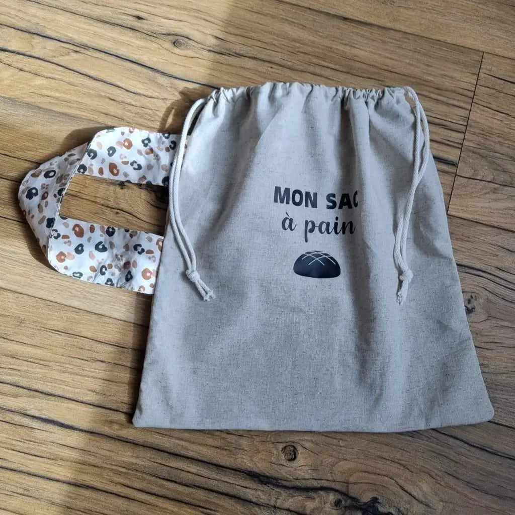 Sac à pain en coton au format miche se fermant par deux petites cordelettes blanches avec une sangle au motif léopard et l'inscription "Mon sac à pain" et le logo d'une miche de pain floqués au centre sur la partie supérieure haute.