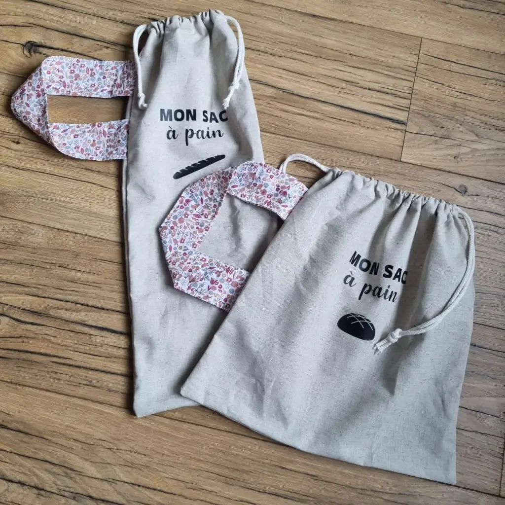 A gauche, un sac à pain en coton au format baguette. A droite, un sac à pain en coton au format miche. Les deux sacs se forment grâce à deux petites cordelettes blanches. La sangle de chaque sac est composée d'un tissu au motif Liberty. Les deux sacs ont l'inscription "Mon sac à pain" floquée au centre sur la partie supérieure haute avec le logo d'une baguette ou d'une miche de pain juste en dessous.