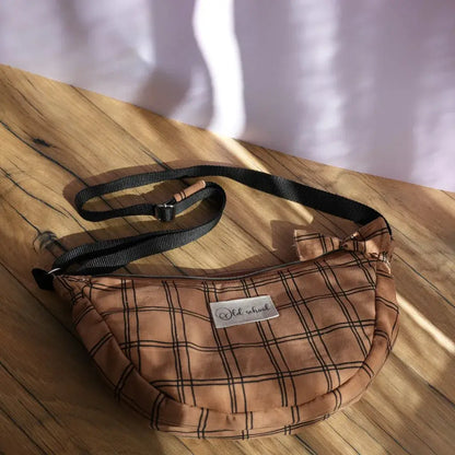 Sac banane demi-lune en tissu marron décoré de motifs à carreaux noirs, posé sur une table en bois éclairée par une lumière naturelle. Équipé d’une bandoulière réglable noire et d’une fermeture zippée, il arbore une étiquette “Old school” cousue sur le devant. Accessoire pratique et tendance, idéal pour transporter l’essentiel tout en ajoutant une touche rétro chic à une tenue décontractée.
