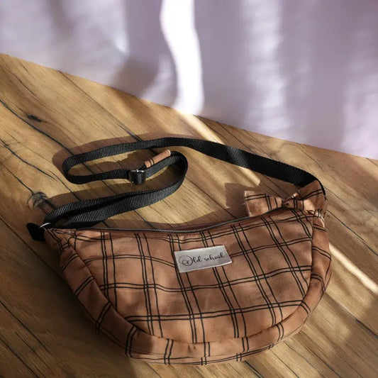 Sac banane demi-lune en tissu marron décoré de motifs à carreaux noirs, posé sur une table en bois éclairée par une lumière naturelle. Équipé d’une bandoulière réglable noire et d’une fermeture zippée, il arbore une étiquette “Old school” cousue sur le devant. Accessoire pratique et tendance, idéal pour transporter l’essentiel tout en ajoutant une touche rétro chic à une tenue décontractée.